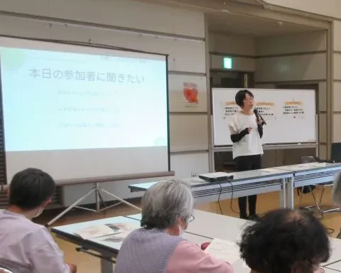 公民館でのセミナー。スクリーンの前で話すいわたまきこと、席につく参加者